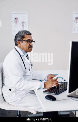 Alles ist in Ordnung, so wie es aussieht. Ein fokussierter, reifer Arzt, der tagsüber an einem Computer arbeitet, während er in seinem Büro sitzt. Stockfoto