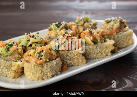 Japanische Tempura-Sushi-Brötchen auf weißem Teller auf Holzhintergrund. Sushi-Stücke mit Lachs, Gurke, Frischkäse, Avocado in knusprigem Nähzeug Stockfoto