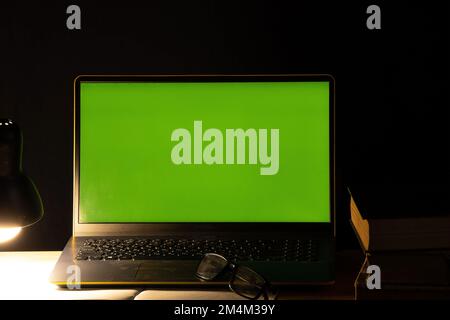 Laptop mit grünem Bildschirm auf dem Tisch mit Büchern in der Nähe der mitgelieferten hellen Lampe im dunklen Schlafzimmer zu Hause Stockfoto