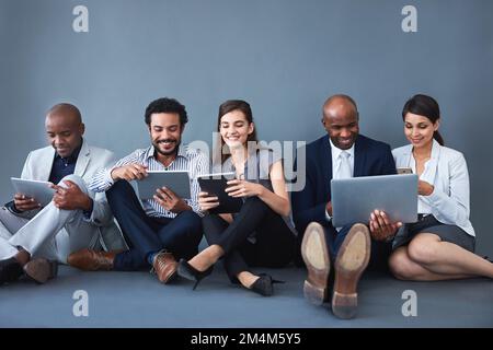 Gute Zusammenarbeit auf verschiedenen Geräten. Studioaufnahme einer Gruppe von Geschäftsleuten mit verschiedenen Geräten vor grauem Hintergrund. Stockfoto