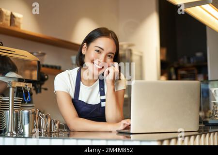 Porträt einer lächelnden koreanischen Frau, eines Baristas im Café, der mit einem Laptop an der Theke steht, lächelt und selbstbewusst aussieht, eine selbstständige Frau Stockfoto