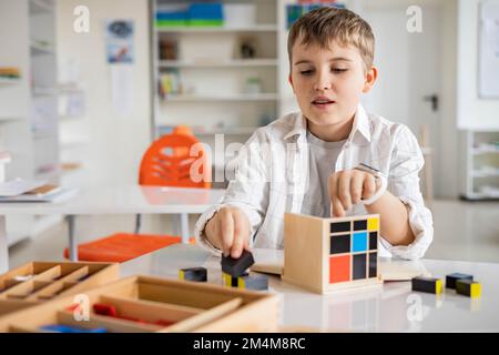 Glücklicher Junge, der Montessori-Stoff-Trinomialwürfel am Klassenschreibtisch zusammenstellt Stockfoto