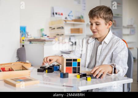 Glücklicher Junge, der Montessori-Stoff-Trinomialwürfel am Klassenschreibtisch zusammenstellt Stockfoto