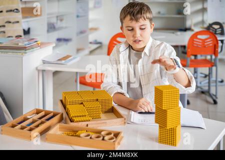 Lächelnder männlicher Junge mit Montessori-Mathematik, goldene Bank, Perlen, alternative Zählentwicklung Stockfoto