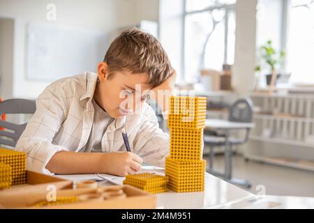 Lächelnder männlicher Junge mit Montessori-Mathematik, goldene Bank, Perlen, alternative Zählentwicklung Stockfoto