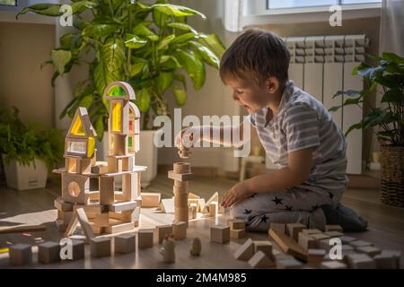 Süßer kleiner Junge, der einen Turm aus Holzziegeln baut, Montessori-Materialien in einem sonnigen Zuhause Stockfoto