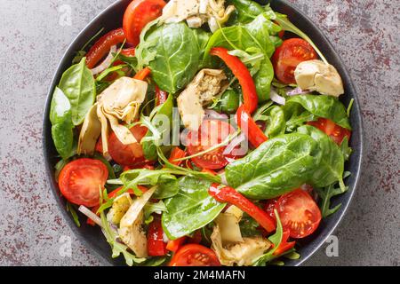 Veganer Diätsalat mit Artischockenherzen, Kirschtomaten, Spinat, Rucola, gebratenen Paprika und Zwiebeln in Nahaufnahme auf einem Teller auf dem Tisch. Horizontale Draufsicht Stockfoto