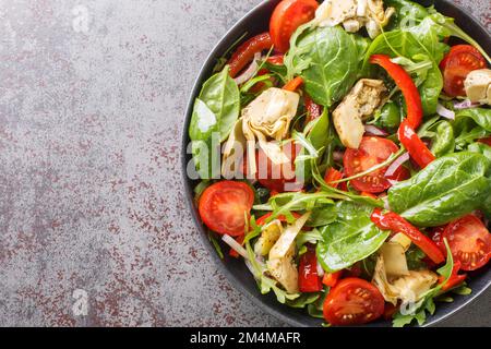 Köstlicher, gesunder Salat aus Artischockenherzen, Tomaten, Spinat, Rucola, gebratenen Paprika und Zwiebeln, Nahaufnahme auf einem Teller auf dem Tisch. Horizontale Draufsicht Stockfoto