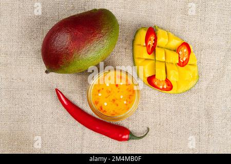 Draufsicht auf ganze Mango, ganze Chilischote, in Scheiben geschnittener scharfer Pfeffer, geschnittene Hälfte der Mango und würziges Mango-Chutney in Glasschüssel auf Sackleinen. Stockfoto