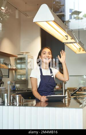 Vertikale Aufnahme eines fröhlichen jungen Baristas, asiatischer Café-Mitarbeiter winkt dem Kunden zu, erhält Bestellungen hinter dem Tresen, verwendet ein Tablet als POS-Terminal und arbeitet in Stockfoto