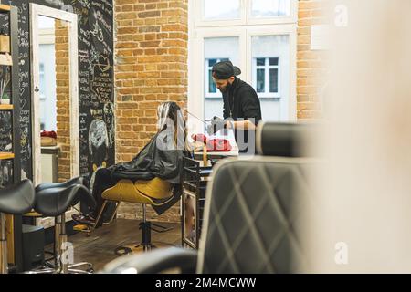Friseurin während der Arbeit. Cline mit langen Haaren sitzt auf einem Stuhl beim Friseur. Durchgehende Aufnahme in der Halle. Friseursalon. Hochwertiges Foto Stockfoto