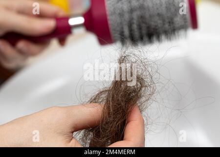 Selektiver Fokus auf die Hand einer Frau mit einem Tropfen fallendes Haar und Bürste im Hintergrund. Zum Problem des massiven Haarverlustes. Haare, Alopezie und Trichot Stockfoto