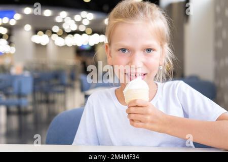 Porträt eines kleinen hungrigen lustigen Mädchens, das in einem Café in einem Waffelbecher kaltes, leckeres Eis isst, dunkler Hintergrund, Bokeh. Ein Kind genießt Eis Stockfoto