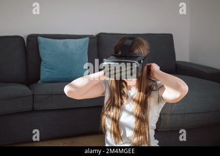 Junge Frau, die mit einem VR-Headset in die virtuelle Welt eintaucht, während sie im gemütlichen Wohnzimmer sitzt Stockfoto