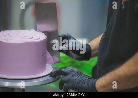 Konditorin mit Spatel und Schaber, glättend, lila Buttercreme, Milchkuchen - Zubereitung von Hochzeitstorten Stockfoto