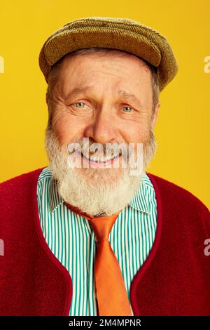 Porträt eines Seniorenmannes in klassischer Kleidung, Brillen- und Mützenposierung, lächelnd auf leuchtend gelbem Hintergrund. Ein lockerer Mann Stockfoto