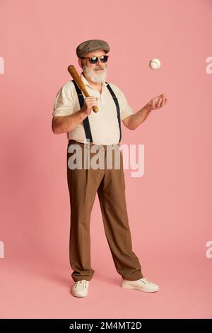 Porträt eines Seniorenmannes in stilvoller Kleidung mit Hosenträgern, Mütze und Sonnenbrille mit Baseballschläger und Ball auf pinkfarbenem Hintergrund. Stockfoto