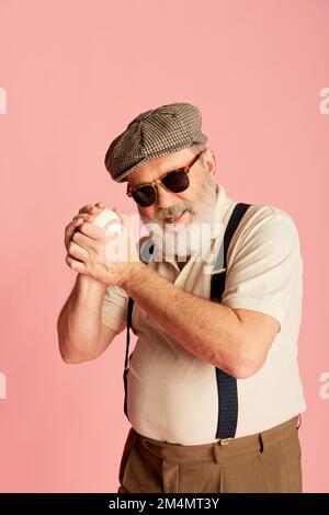 Porträt eines Seniorenmannes in stilvoller Kleidung, Mütze, Hosenträgern und trendiger Sonnenbrille mit Baseballball auf pinkfarbenem Hintergrund. Stockfoto