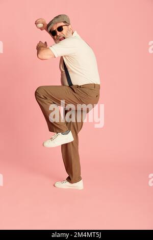 Porträt eines Seniorenmannes in stilvoller klassischer Kleidung, Posen, Servieren eines Baseballballs auf pinkfarbenem Hintergrund. Sportverein Stockfoto