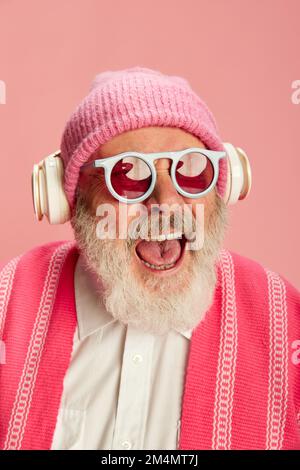 Porträt eines stilvollen älteren Mannes in stilvollem, hellen Outfit mit Strickmütze und Sonnenbrille, die in Kopfhörern auf pinkfarbenem Hintergrund posieren Stockfoto