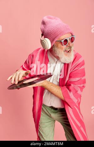Gefühlvoller alter Mann in elegantem, hellen Outfit mit Strickmütze, Sonnenbrille, Kopfhörern und Vinyl auf pinkfarbenem Hintergrund. Dj Stockfoto