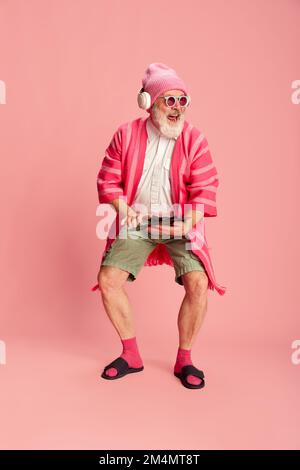Porträt eines stilvollen älteren Mannes in elegantem, hellen Outfit mit Sonnenbrille und Kopfhörern auf pinkfarbenem Hintergrund. Hipster Stockfoto