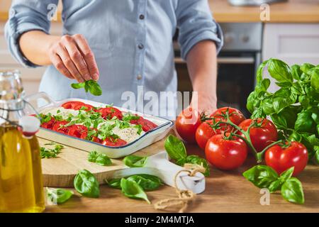 Ernten Sie anonyme Hausfrau in blauem Hemd und servieren Sie Caprese in quadratischer Keramikschale, während Sie neben einer Holztheke mit frischen Tomaten und einer Flasche Öl stehen Stockfoto
