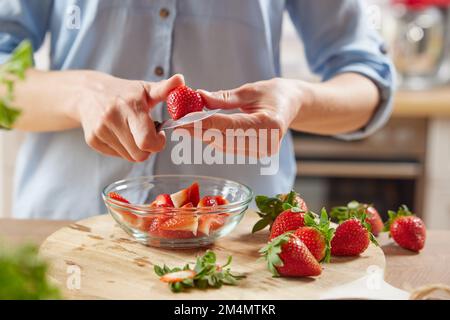 Schneiden Sie eine anonyme Frau frische Erdbeeren in eine Glasschale, während Sie ein gesundes Frühstück zuhause in der Küche zubereiten Stockfoto
