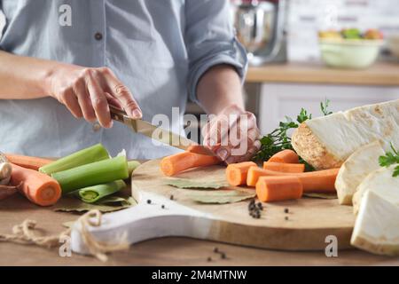 Ernten Sie eine anonyme Frau, die frische Karotten mit dem Messer auf Holzbrett schneidet, während Sie gesunde Lebensmittel zuhause in der Küche zubereiten Stockfoto