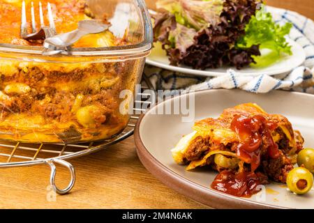 Ein Stück köstliche hausgemachte Lasagne in brauner Keramikschale mit Lasagne in transparenter Glasschale auf einem Kühlregal und frisches Gemüse auf einer weißen Platte Stockfoto