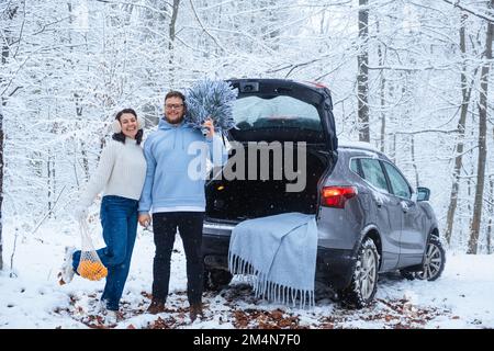 Ein glückliches Paar, das sich in der Nähe des Kofferraums im verschneiten Wald verliebt Stockfoto