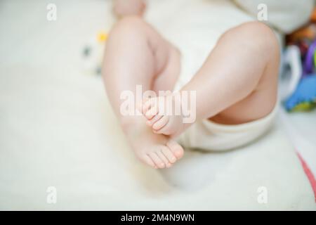 Gekreuzte Beine des Babys auf weißem Hintergrund. Ein kleines Mädchen liegt auf einem weißen Bett in einem weißen Body. Stockfoto