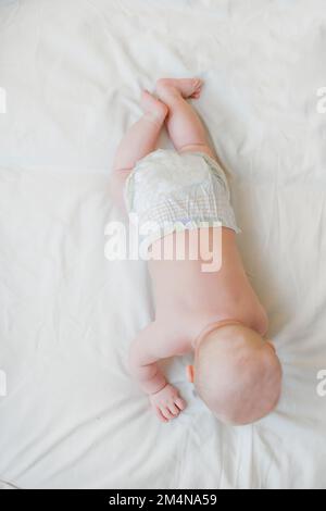 Das Baby liegt auf dem Bauch auf einem weißen Laken auf dem Bett von oben. Das Neugeborene in der Windel ruht vor dem Schlafengehen Stockfoto