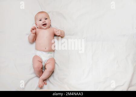 Ein lächelndes Baby liegt auf einem weißen Bett. Ein kleines Kind in der Windel freut sich über die Ankunft seiner Mutter und lächelt. Stockfoto