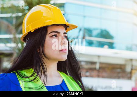 Porträt einer jungen Kaukasierin, blau gekleidet, mit gelbem Schutzhelm und Sicherheitsweste, die draußen steht und in die Distanz schaut Stockfoto