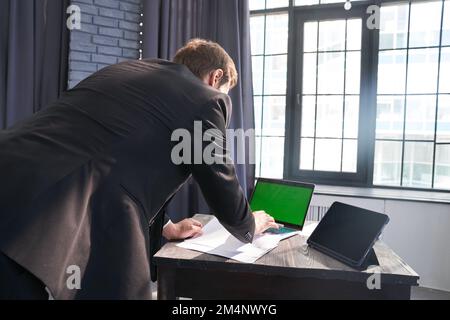 Ein unbekannter Geschäftsmann, steht und tippt auf seinem Laptop mit einem Chromakey auf dem Hintergrund des Fensters. Mit einem Platz zum Kopieren. Hochwertiges Foto Stockfoto