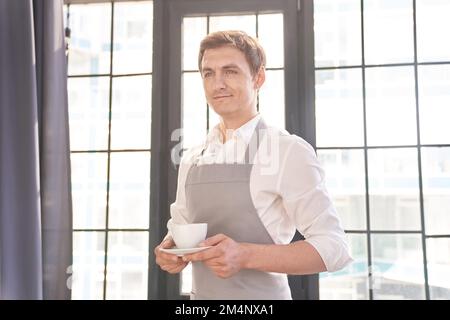 Ein männlicher Kellner blickt in einer grauen Schürze weg und hält eine Tasse Kaffee aus. Der Barista serviert eine Tasse heißen Kaffee vor dem Hintergrund eines großen Fensters. Hochwertiges Foto Stockfoto