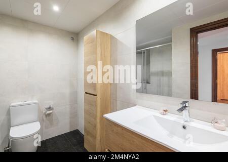 Modernes Bad mit weiß gefliesten Wänden. Der kleine Holzschrank für Toilettenartikel und Handtücher sitzt an der Wand und harmoniert farblich mit dem Schrank für Waschbecken Stockfoto