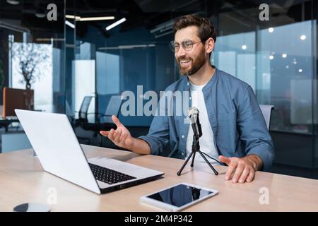 Ein lächelnder junger Mann mit Brille sitzt im Büro am Tisch und spricht bei einem Videoanruf von einem Laptop in ein Mikrofon. Schreibt einen Blog, Podcast, ein Interview, führt ein Webinar durch, führt Geschäftsschulungen durch. Stockfoto