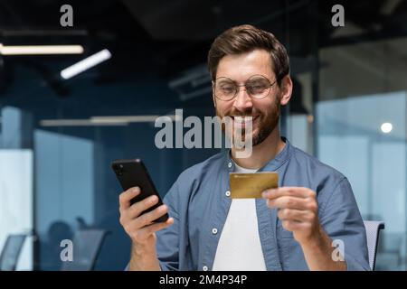 Ein lächelnder junger Mann hält ein Telefon und eine Kreditkarte. Steht im Büro, wählt am Telefon, tätigt Online-Käufe, bestellt, prüft das Konto. Stockfoto