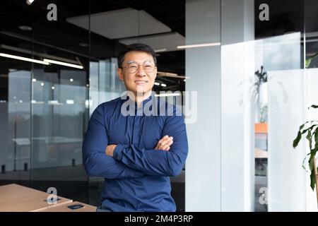 Portrait eines erfolgreichen lächelnden Geschäftsmanns, eines Asiaten mit zerknitterten Händen in einem legeren Hemd, der aus dem Fenster schaut, eines erwachsenen Mannes mit Brille, der in einem modernen Büro arbeitet. Stockfoto