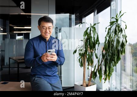 Ein asiatischer Geschäftsmann im Büro in einem lässigen Hemd und einer Brille benutzt das Telefon, der Chef hält ein Smartphone in den Händen, liest Nachrichten und tippt lächelnd eine Nachricht. Stockfoto