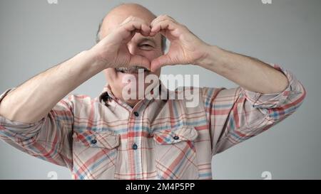Ein hübscher, verliebt lächelnder Senior-Mann, der ein Herzsymbol mit Händen formt. Romantisches Konzept. Stockfoto