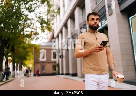 Blick auf einen selbstbewussten jungen Mann mit tätowierter Hand, der auf der Straße spaziert, am Sommertag Smartphone benutzt und Kaffee zum Mitnehmen trinkt. Stockfoto