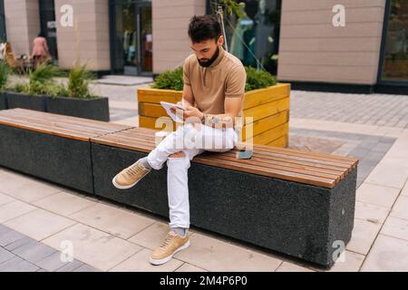 Weitwinkelaufnahme eines talentierten männlichen Künstlers mit tätowierten Händen, die auf der Bank sitzen, Kaffee zum Mitnehmen trinken und mit Stift auf Copybook Skizzen machen. Stockfoto