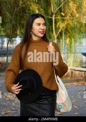 Attraktive, stilvolle Frau, die Hut und Tasche in der Hand hält, im Park spaziert, in einem warmen Anzug gekleidet. Hübsche Frau mit braunen Haaren lächelt breit die Zähne. Herbst Stockfoto