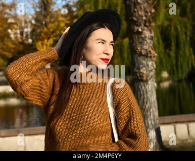 Selektiver Fokus einer lächelnden, modischen jungen gebräunten Frau mit Hut auf der Herbstlandschaft im Herbst im Stadtpark. Positives Emotionskonzept. Stockfoto