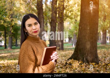 Seitenansicht einer charmanten Frau, die im Gras unter dem Baum ein Buch liest und eine Tasse Kaffee trinkt. Entspannen Sie sich im Sommerurlaub bequem auf dem Rasenplatz Stockfoto