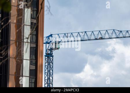 Blauer hoher Kran auf der Baustelle mit bewölktem, malerischem Himmel in der Nähe eines modernen Gebäudes mit brauner und weißer reflektierender Glasfassade Stockfoto