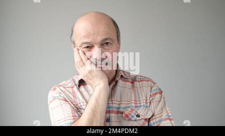 Ein sehr gelangweilter älterer Erwachsener, der die Hand auf der Wange hält, sieht müde und krank aus, mit grauem Hintergrund. Großvater wartet auf seine Familie Stockfoto